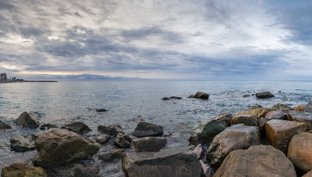 Surf waves of sea between the coastal rocks of a stone coastline and water surface against the cloudy sky, panoramic view in the autumn overcast morningの写真素材