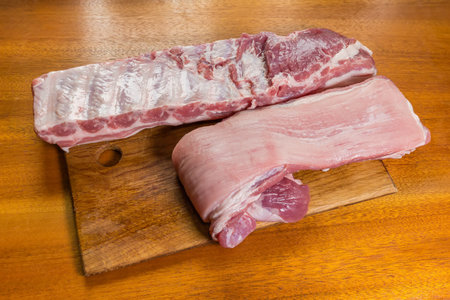 Two large whole pieces of the raw chilled pork ribs in the form of long strips lie on a wooden cutting board on a wooden tableの写真素材
