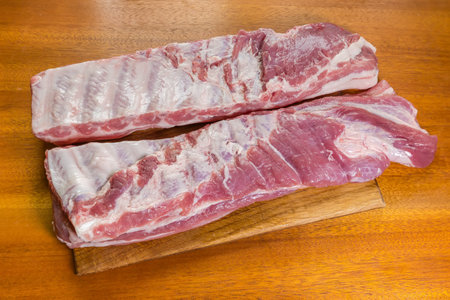 Two large whole pieces of the raw chilled pork ribs in the form of long strips lie on a wooden cutting board on a wooden tableの写真素材
