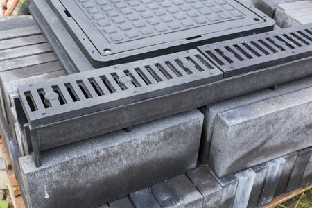 Plastic gutter of the rain drain with drainage grates atop lies on a stack of other construction materials on a background of the square sewer hatches outdoorsの写真素材