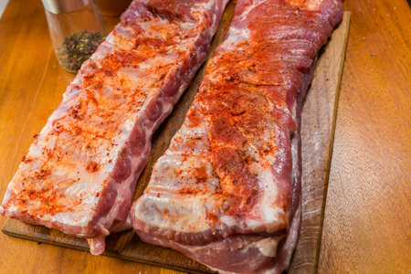 Two whole pieces of the raw pork ribs sprinkled with various spices lie on a wooden cutting board on a wooden table, close-up in selective focusの写真素材