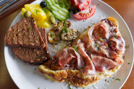 Omelette served with grilled bacon and mushrooms, olives and fresh vegetables, sprinkled with grated cheese and chopped greens, toasts of brown bread, top view in selective focusの写真素材