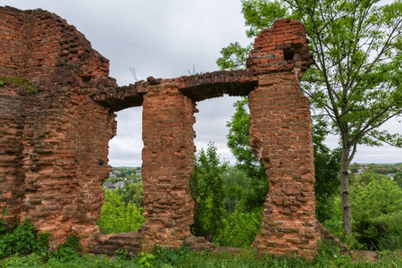 Fragment of the remains of defense red brick wall with two arched window openings of ruined medieval castle in spring overcast day, view through themの写真素材