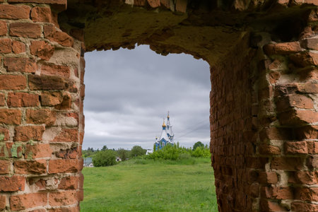 View of the distant old wooden church through the window opening of the defense red brick wall in ruined medieval castle in spring overcast dayの写真素材