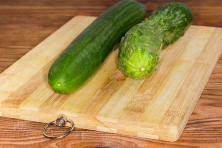 Two whole fresh green long cucumbers of different varieties with smooth and bumpy skin on a wooden cutting board on a rustic table, front view close-up in selective focusの写真素材