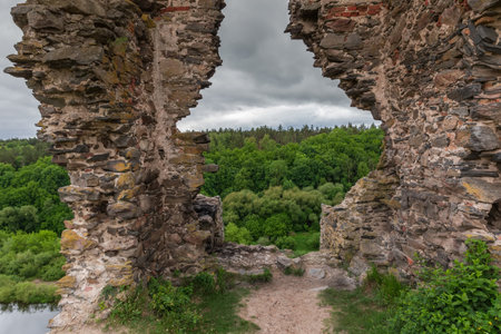 Part of ruins of the stone defensive tower of Hubkiv Castle of the 15th century in overcast day, Ukraine, view of forest on opposite bank through a hole in the wallの写真素材
