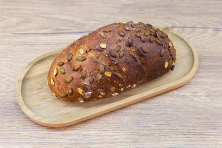 Whole oblong loaf of grain bread sprinkled with pumpkin seeds on the wooden tray on a wooden table