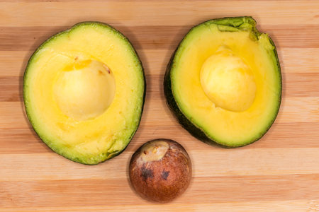 Two halves of the ripe avocado with dark green skin and separated stone on a wooden bamboo cutting...