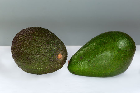 One whole ripe green pear-shaped ordinary avocado and one  avocado Hass variety with bumpy purplish-black skin on a white surface, front view on a gray backgroundの写真素材