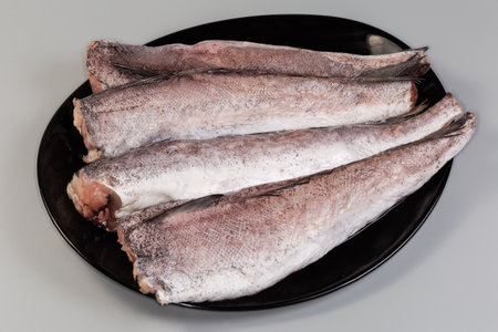 Uncooked defrosted carcasses of European hake without the heads and tails on a black plate on a gray backgroundの写真素材