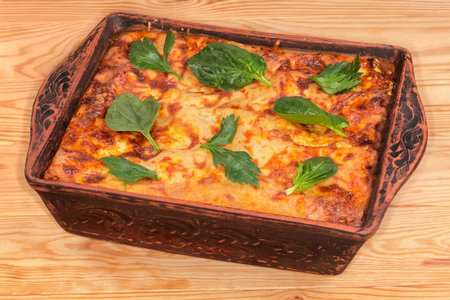 Baked potato casserole with browned crust of the melted cheese on top and decorated with fresh celery and spinach leaves in the rectangular clay oven tray on a rustic tableの写真素材