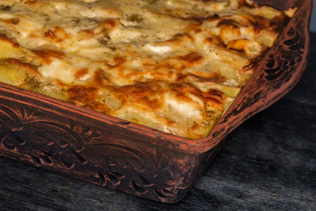 Baked potato casserole with browned crust of the melted cheese on top in the rectangular clay oven tray on a black wooden table, fragment close-upの写真素材