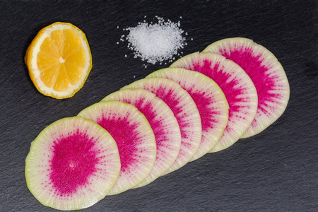 Round thin slices of the watermelon radish, fresh lemon piece and salt on a black flat slate plate,...