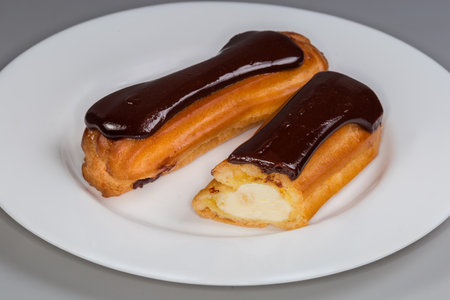 One whole and one halved eclairs with custard and chocolate glaze on a white plate, front view...