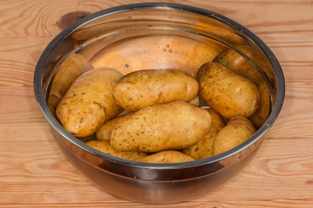 Raw washed yellow oblong potato tubers in their skins in a stainless steel kitchen bowl on a wooden rustic tableの写真素材