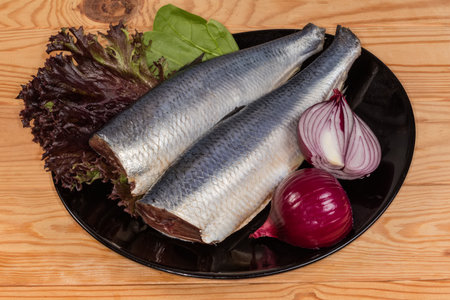 Two whole pickled gutted atlantic herrings without heads and tails with onion and leaf vegetables on...