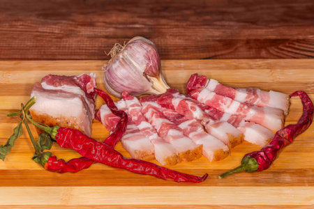 Piece and slices of the salted pork belly with significant amount of the lean meat streaks, on a skin, dry chili pods and garlic on a bamboo cutting board, front view close-up in selective focusの写真素材
