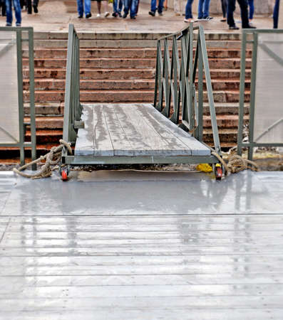 Wooden gangway between  coast and  floating mooring. On  embankment blur  legs of the people...