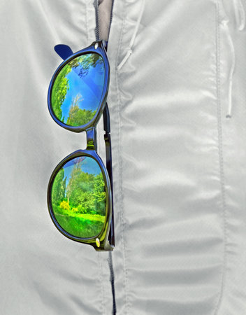 Sunglasses hang on zipper of white jacket . Lenses and frame reflect blur bright spring landscape.の写真素材