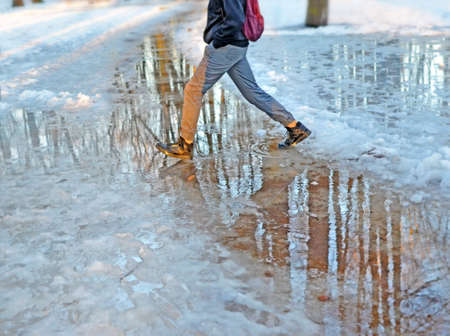 Legs of young man who walks through park in the early spring and steps over puddle. Splashes darken on his pants and shine in sun on his shoes. Shadow of tree falls on his body. Meltwater reflects blue sky and bare trunks.の写真素材