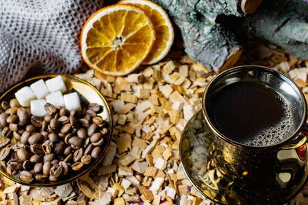 Aromatic dark coffee with bubbles on the surface in a dark gold Cup on a saucer, next to whole grains, peeled and dried oranges. Background of alder chips and in the background snags of wood with barkの写真素材