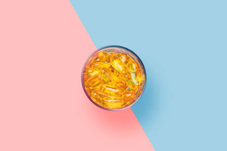 Bright yellow fish oil capsules in the glass cup on bright pink and blue background shot with studio lightの写真素材