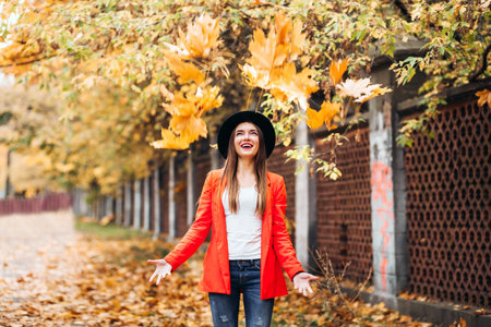 Happy young woman in red jacket and black hat throwing leaves and smiling on colorful nature...