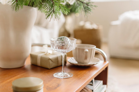 Cozy winter home decor scene with a crystal glass holding a shiny Christmas ornament, a cup of coffee, and a wrapped gift on a wooden table. Warm festive atmosphere with elegant minimalist details and soft natural light.の写真素材