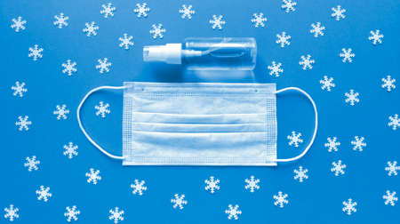 White medical mask and hand sanitizer in transparent bottle with spray cap at the middle of blue background and scattered white snowflakes. Simple festive flat lay. Medical concept. Stock photo.の写真素材