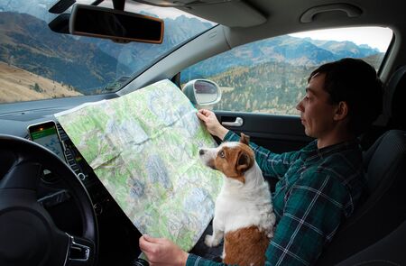 young man with a dog in a car are watching a map in the mountains. Hipster, Hiking concept,...