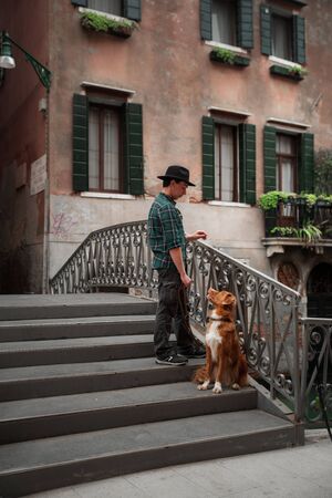 A man with a dog on a waterfront in Venice. Traveling with a pet. Nova Scotia Duck Tolling Retriever on a leash with the cityの写真素材