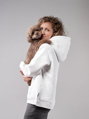 happy girl with a dog in a photo studio on gray. Curly woman in a white hoodie with a chocolate poodle in her arms.の写真素材