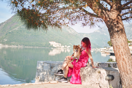 A woman with pink hair sits beside a Shiba Inu dog, both gazing out at a lake