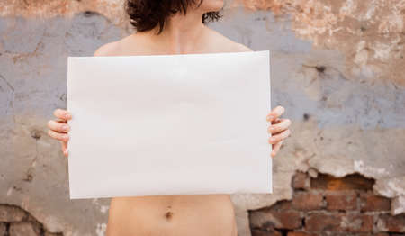A girl stands with an empty poster against the destroyed wall of a building. Single picket of a woman on the protection of rights with a blank sheet of paperの写真素材