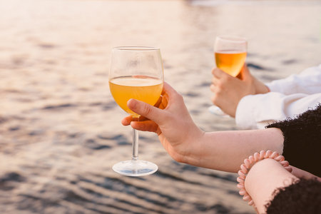 glasses with cider in the hands of people on the background of the water surface in the sunset lightの写真素材
