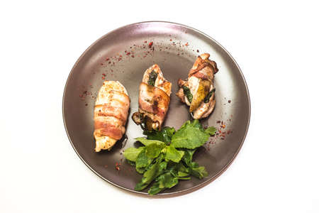 White chicken meat in bacon with greens on a black plate on a white backgroundの写真素材
