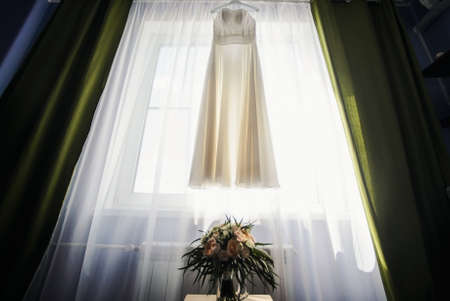 beautiful white luxury wedding dress on hanger on the background of a window with flowersの写真素材