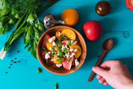 Making a salad of freshly colored tomatoes. Salad of slices of tomato, cheese and green onions on a bright turquoise background and a male hand. View from above.の写真素材