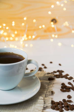 Black coffee in a white cup and whole grains of coffee on a background of defocused lights of a garland. Creative coffee concept. Coffee drink winter holiday new year.の写真素材