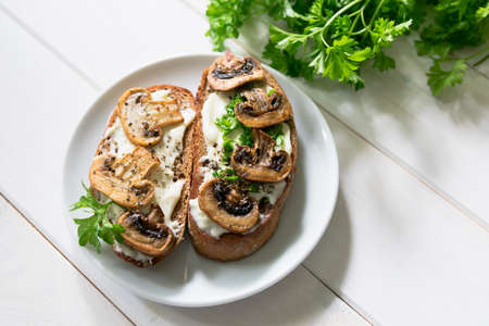 Two sandwiches with mushroom slices and curd cheese and fresh bright parsley lie on a white plate on a white background.の写真素材