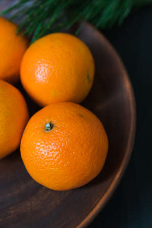 Fresh tangerines lie on an old clay plate close-up, dark background and coniferous tree branch. Rustic concept. Holidays, New Year, Christmas. Winter still lifeの写真素材