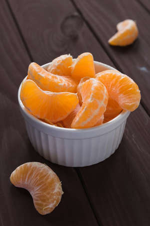 Slices of ripe mandarin lie in a white bowl next to another slices on a dark brown background.の写真素材