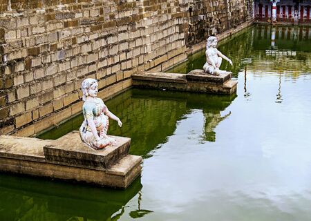 Two ancient statues - a woman and a man, on ledges in the wall of an old pondnear Indian templeの写真素材