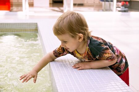 Little blond boy with joy and curiosity touches by hand the water in the fountain  in shopping...