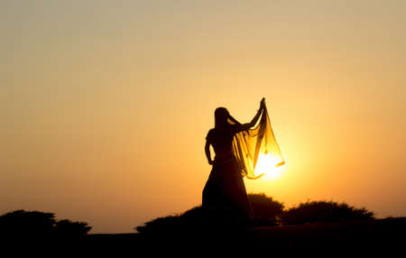 Young woman in Indian clothes dances at sunset in the desert on the dunes against low bushes, sun and sky. Rajasthan, India.の写真素材