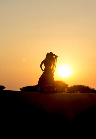 Young woman in Indian clothes dances at sunset in the desert on the dunes against low bushes, sun and sky. Rajasthan, India.の写真素材