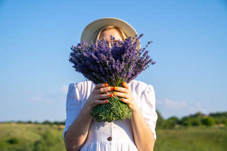 portrait of woman model in white dress outfit with hat is standing in lavender field, photo session. Young girl is holding lavender bouquet. Good picture for cover, calendarの写真素材