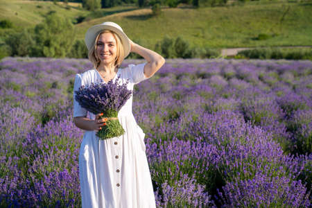 portrait of woman model in white dress outfit with hat is standing in lavender field, photo session. Young girl is holding lavender bouquet. Good picture for cover, calendarの写真素材