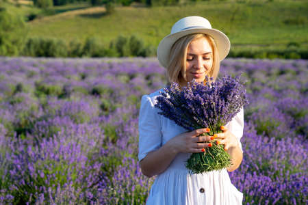 portrait of woman model in white dress outfit with hat is standing in lavender field, photo session. Young girl is holding lavender bouquet. Good picture for cover, calendarの写真素材