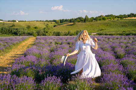 woman model in white dress outfit with hat is standing dancing in lavender field, photo session. Concept of freedom. Good picture for cover, calendar, postcard, wallpaperの写真素材